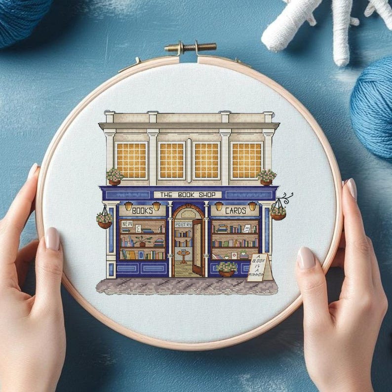 Pode incluir: Bordado em ponto cruz de uma livraria. A fachada da loja &eacute; azul e branca, com a inscri&ccedil;&atilde;o "THE BOOK SHOP" acima da porta. A loja vende "BOOKS" e "CARDS". O bordado &eacute; apresentado num bastidor de madeira.