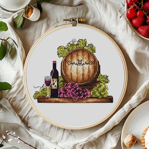 May include: A cross-stitch embroidery design featuring a wooden wine barrel with grapes, a bottle of wine, and a glass of wine. The design is framed in a wooden hoop.