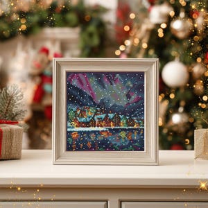 Puede incluir: Un punto de cruz enmarcado que representa una escena de un pueblo invernal bajo una colorida aurora boreal. La obra de arte presenta un paisaje nevado con casas reflejadas en el agua. El marco es de color claro y neutro, y el tema general es festivo.