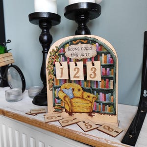 May include: An arched wooden sign featuring a cross-stitch pattern of a bookshelf and a yellow armchair. The text reads "Books read this year" above three hanging wooden number tiles showing 1, 2, and 3, with additional number tiles scattered below.