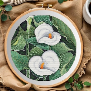 May include: A circular wooden embroidery hoop containing a cross-stitch pattern of two white calla lilies with orange centers, surrounded by large, detailed green leaves against a dark, textured background.