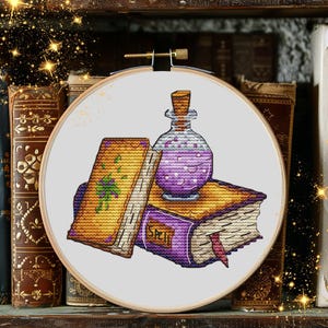 May include: A cross-stitch embroidery in a circular wooden hoop featuring a yellow book with a green design, a purple book labeled SPELL, and a glass bottle filled with a purple liquid. The design is stitched on white fabric with detailed thread work.