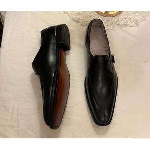 May include: A pair of black leather dress shoes with a single buckle strap. The shoes have a pointed toe and a brown leather sole.