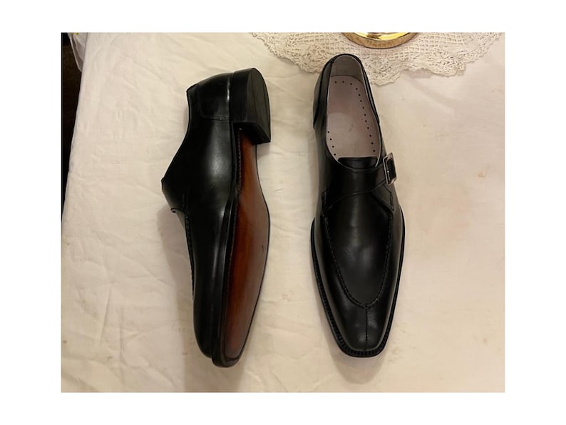 May include: A pair of black leather dress shoes with a single buckle strap. The shoes have a pointed toe and a brown leather sole.
