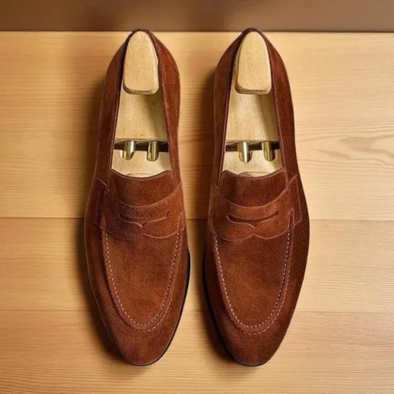 Puede incluir: Un par de mocasines penny de ante marr&oacute;n con hormas de madera. Los zapatos tienen un dise&ntilde;o cl&aacute;sico con punta redondeada y una correa en el empeine. Se exhiben sobre una superficie de madera marr&oacute;n claro.