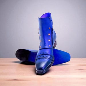 May include: A pair of blue leather ankle boots with copper-colored studs and a side zipper. The boots have a pointed toe and a stacked heel. The boots are displayed on a wooden surface.