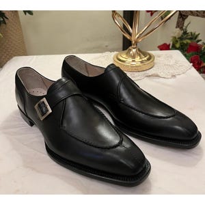 May include: A pair of black leather dress shoes with a single monk strap buckle. The shoes have a pointed toe and a slightly raised heel.