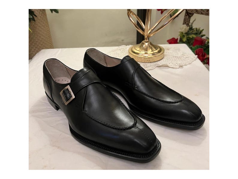 May include: A pair of black leather dress shoes with a single monk strap buckle. The shoes have a pointed toe and a slightly raised heel.