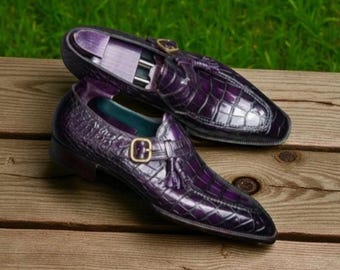 May include: A pair of purple leather loafers with a crocodile skin texture. The shoes have a gold buckle and tassel detail. The loafers are displayed on a wooden surface with a green grass background.