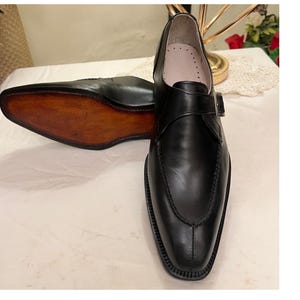 May include: Black leather dress shoes with a single buckle strap. The shoes have a pointed toe and a brown leather sole.
