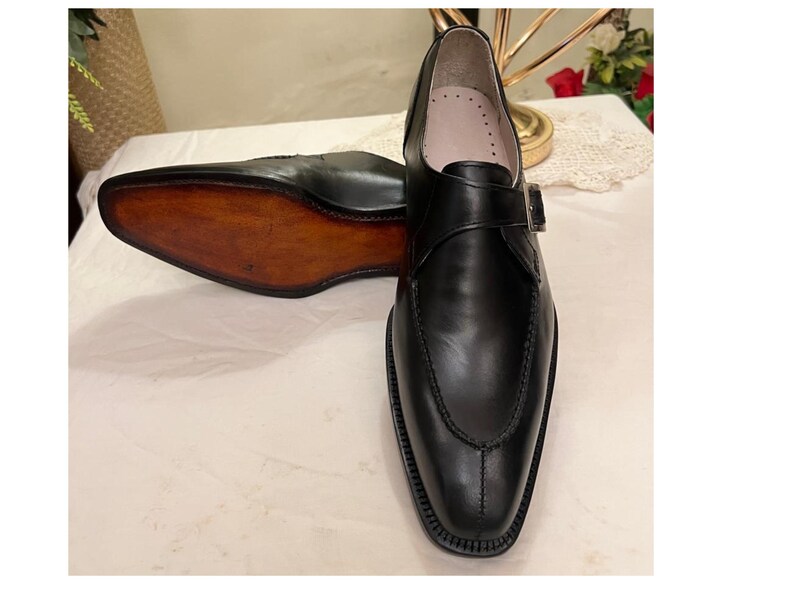 May include: Black leather dress shoes with a single buckle strap. The shoes have a pointed toe and a brown leather sole.