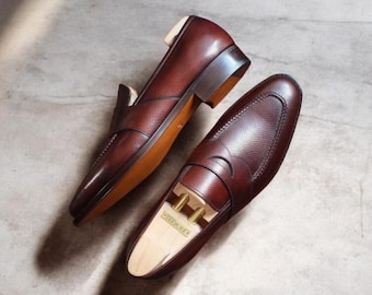 May include: A pair of brown leather loafers with a textured vamp and a smooth, glossy finish. The shoes have a classic design with a low heel and a slip-on style. The image shows the shoes on a neutral surface.