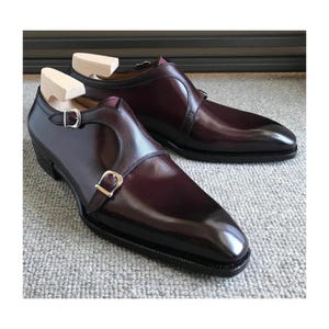 Pode incluir: Um par de sapatos de couro bordô com fivelas duplas. Os sapatos têm bico fino e acabamento polido.
