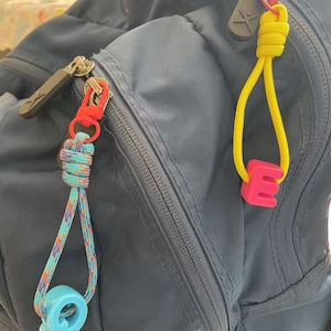 May include: A navy blue backpack with two custom zipper pulls. One has a red clasp and a turquoise and orange rope with a light blue letter "Q". The other has a yellow rope and a pink letter "E".