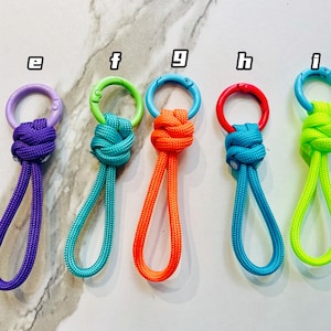 May include: Five brightly coloured paracord keychains with metal rings. The keychains are in purple, teal, orange, blue, and neon green. Each keychain features a knotted design and a loop for easy attachment.