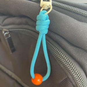 Op de afbeelding: Een close-up van een turquoise paracord ritstrekker met een oranje kraal. Het paracord is geknoopt en bevestigd aan een zwarte tasrits. De kraal is fel oranje.