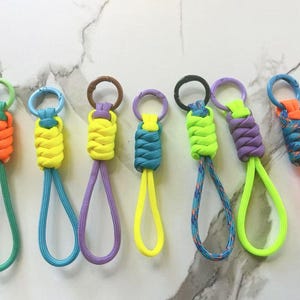 May include: A collection of colorful paracord keychains. Each keychain features a woven knot design in various colors, including orange, yellow, teal, purple, and neon green. The keychains have a loop at the bottom and a metal ring at the top.