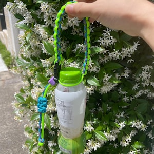 May include: A clear plastic bottle with a green cap is held by a neon green and blue braided strap. The strap has a black buckle and a teal and purple accent. The bottle is filled with a white liquid and has a green label.