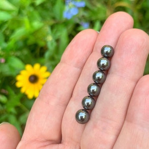 May include: Six small, dark gray, spherical beads are arranged in a vertical line, held in an open hand. The beads have a shiny, reflective surface. The background is a blurred view of green foliage and a yellow flower.