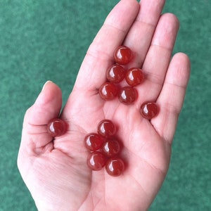 Può includere: Una mano che tiene una collezione di 12 piccole perle rotonde di cornalina rossa.