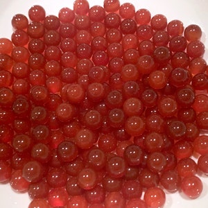 May include: A close-up of a bowl filled with small, round, translucent red beads. The beads are arranged in a random pattern and appear to be made of glass or plastic.