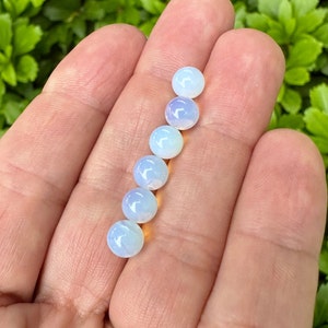 May include: A row of seven iridescent white opal beads, each with a slightly different shade of blue and white, are displayed on a hand.