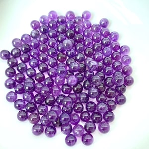 May include: A collection of polished, round amethyst gemstones in varying shades of purple. The gemstones are scattered on a white surface, with each stone reflecting light. The overall effect is a display of natural beauty and color.