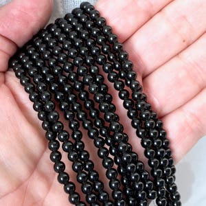 May include: A strand of black onyx beads, approximately 4mm in diameter, displayed on a hand. The beads are smooth and polished, with a glossy finish.