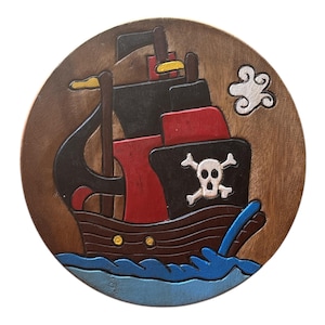 Könnte beinhalten: Ein hölzerner Piratenschiffschnitzerei auf einer runden Holzbasis. Das Schiff hat schwarze Segel, einen roten Rumpf und ein Totenkopf-Emblem. Blaue Wellen und eine weiße Wolke vervollständigen das Design.