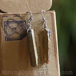 May include: A pair of gold-toned earrings with long, rectangular, crystal-like pendants. The earrings are hanging from silver hooks.
