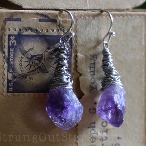May include: Amethyst crystal earrings with silver wire wrapping. The amethyst crystals are a deep purple color and have a rough, natural texture. The earrings are hanging from silver hooks.