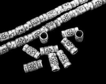 May include: Silver-colored metal beads and bead components. The beads are cylindrical with a repeating cross and diamond pattern. Some beads are strung together, while others are loose. The background is black.