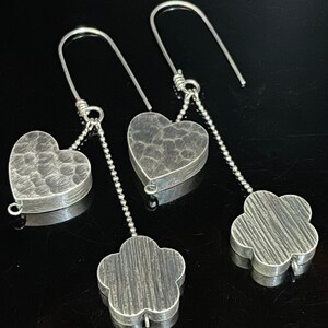 May include: A pair of silver dangle earrings with a heart and flower charm. The charms have a hammered texture.