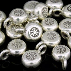 May include: A collection of silver-colored metal beads. Each bead is round with a loop and a sunburst design on the top. The beads are scattered on a dark surface, creating a textured appearance. Ideal for jewelry making.