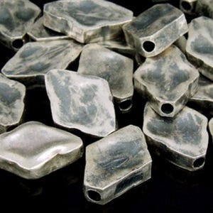 Op de afbeelding: Een stapel zilverkleurige, onregelmatig gevormde, diamantvormige kralen met een gat in het midden van elke kraal.