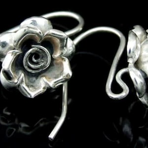 May include: A pair of silver-toned rose earrings. Each earring features a detailed rose blossom with a spiral center and a curved ear wire. The earrings are set against a black background, highlighting their metallic sheen.
