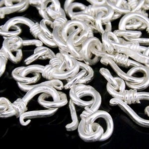 May include: A close-up of a pile of silver-toned metal clasps. The clasps are intricately designed with a twisted wire detail and a hook shape.