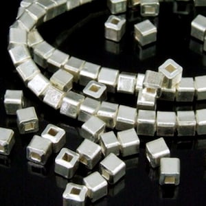 May include: Silver square beads with a hole in the center. The beads are arranged in a circular pattern and are scattered on a black surface.