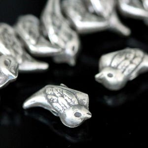 Könnte beinhalten: Eine Gruppe von silbernen Vogelanhängern, jeder mit detaillierten Flügeln und einem kleinen Schnabel. Die Anhänger sind in einer Reihe angeordnet, wobei sich einige überlappen.