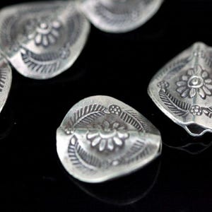 Pode incluir: Contas de metal prateadas com um design floral. Cada conta tem uma flor em relevo, detalhada, no centro, cercada por folhas. As contas têm a forma de uma lágrima.