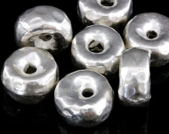 Può includere: Un gruppo di perline in metallo color argento con una texture martellata e un foro rotondo al centro. Le perline sono disposte in modo casuale.