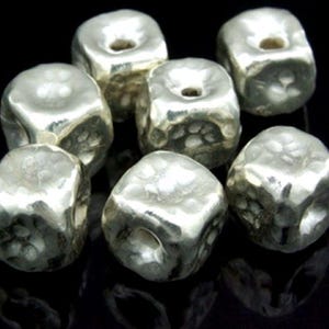 May include: A group of eight silver-toned metal cube beads with a hammered texture. The beads have a hole in the centre of each side.