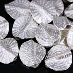 May include: A group of silver-colored metal beads with a textured, leaf-like design. The beads are arranged in a pile, with some overlapping.
