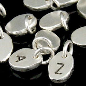 May include: A collection of silver oval charms with a hammered texture. Each charm has a loop for attaching to a necklace or bracelet. Some charms have letters engraved on them, including the letters A and Z.