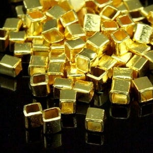 May include: A pile of gold-coloured metal square beads with a hole in the centre. The beads are small and have a shiny finish.