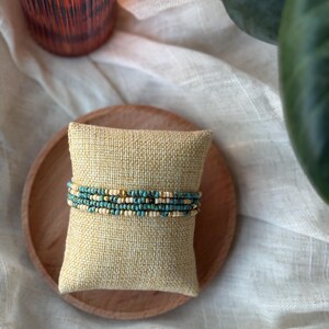 May include: A stack of beaded bracelets in shades of turquoise, cream, and gold, resting on a small, square, beige burlap pillow. The pillow sits on a round, light brown wooden plate, with a white cloth in the background.
