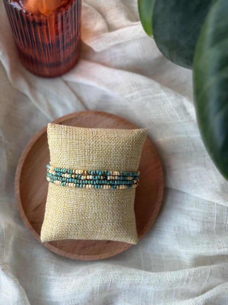 May include: A stack of beaded bracelets in shades of turquoise, cream, and gold, resting on a small, square, beige burlap pillow. The pillow sits on a round, light brown wooden plate, with a white cloth in the background.
