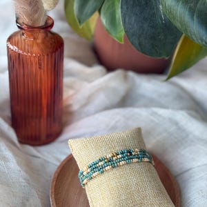 May include: A beaded bracelet with turquoise, cream, and gold beads rests on a small, square, beige pillow. The pillow sits on a round, wooden tray. A textured, orange glass vase with dried flowers and a potted plant are in the background.