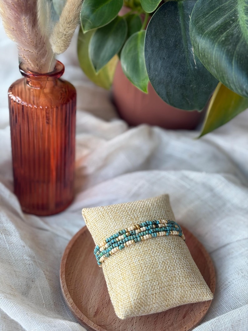 May include: A beaded bracelet with turquoise, cream, and gold beads rests on a small, square, beige pillow. The pillow sits on a round, wooden tray. A textured, orange glass vase with dried flowers and a potted plant are in the background.