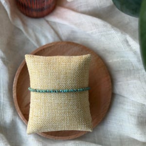 May include: A delicate turquoise beaded bracelet rests on a small, textured, yellow pillow. The pillow sits on a wooden plate, with a blurred background of a glass and greenery. The bracelet is a simple, elegant piece of jewelry.